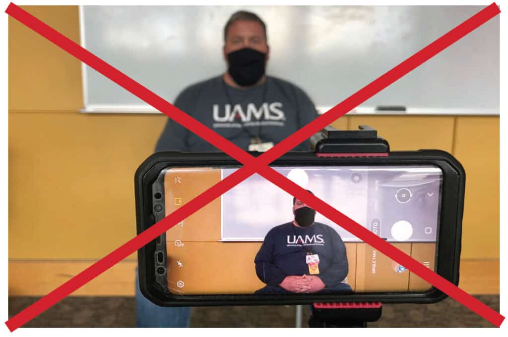 Image has a red X over it and shows a poor example of a video setup. Male is sitting in front of a white dry erase board and through the camera, there are reflections on the board from lights in the room.